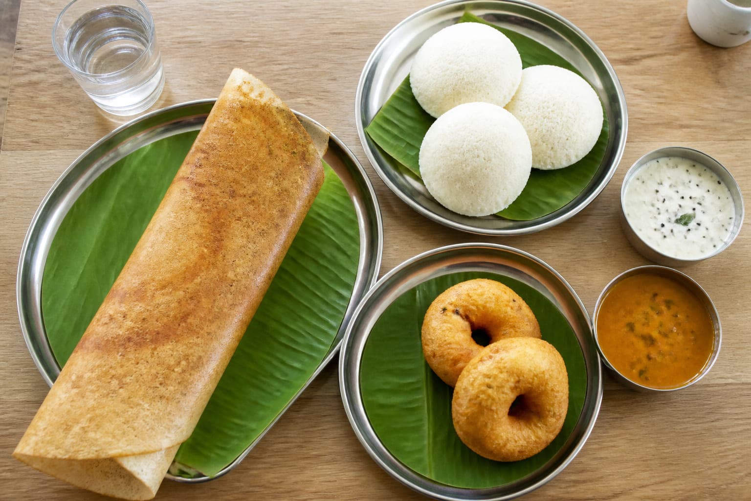 Frühstücksgerichte: eine gerollte Dosa, drei Idli auf Bananenblatt, zwei Vada und zwei Schälchen Chutney